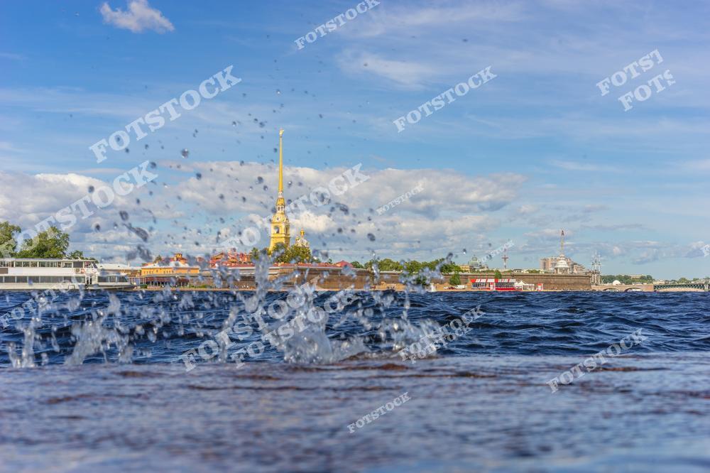 На фотографии изображена Петропавловская крепость с её знаменитым шпилем, отражающимся в воде Невы. Передний план украшен брызгами воды, создающими динамику и свежесть композиции. Ясное небо и солнечный свет подчеркивают красоту летнего дня в Санкт-Петербурге. Эта сцена прекрасно передает атмосферу города, связанного с водой.